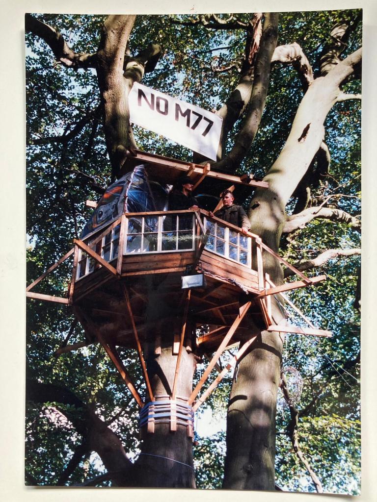 A well-constructed tree house amidst lush green leaves, with two people gazing out from a platform and a banner above them reading NO M77