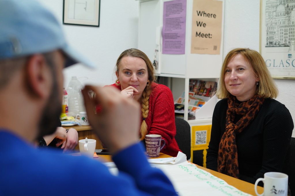 A group of people sit round a table. Two women both with blonde hair are in focus. 